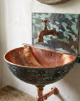 Copper sink with green patina and brass faucet against a white wall.
