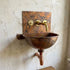 A rustic copper wall-mounted sink with a hammered finish, featuring a matching copper backplate, brass faucet, and wall-mounted pipes against a textured plaster wall.
