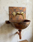 A rustic copper wall-mounted sink with a hammered finish, featuring a matching copper backplate, brass faucet, and wall-mounted pipes against a textured plaster wall.